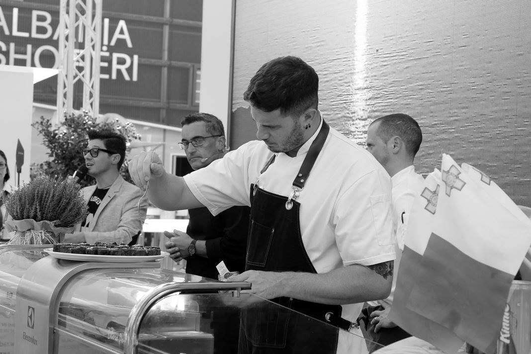 Sean Gravina plating at a live cooking event beside Maltese flags